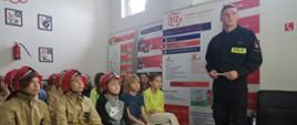 Na zdjęciu widzimy dzieci w strojach strażackich oraz strażak w sali edukacyjnej podczas oglądania filmu