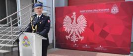 Zdjęcie przedstawia Zastępcę Wielkopolskiego Komendanta Wojewódzkiego PSP, który stoi przy mównicy na placu przed budynkiem komendy podczas uroczystych obchodów dnia strażaka.