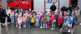 Na zdjęciu widoczna jest duża grupa dzieci z przedszkola, stojących przed czerwonym wozem strażackim. Obok dzieci i wozu strażackiego stoją dorośli, w tym kilku strażaków w mundurach. Na wozie strażackim widać numer 701-21 oraz numery alarmowe 998 i 112. Zdjęcie zrobione podczas wycieczki edukacyjnej w jednostce straży pożarnej, gdzie dzieci mają okazję poznać sprzęt i zawód strażaka.