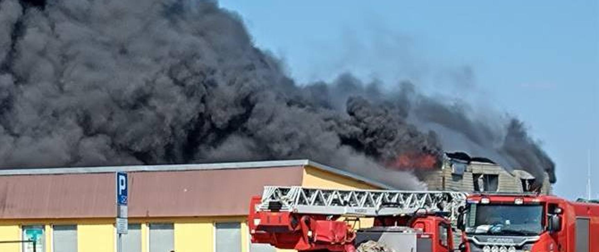 Gęsty dym unoszący się na dachami budynków, na pierwszym planie dwa samochody strażackie i strażak