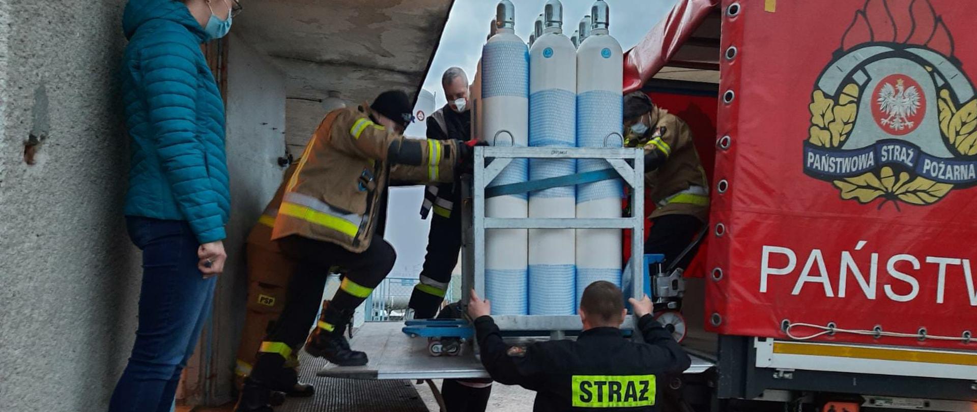 Na zdjęciu z prawej strony samochód Komendy Wojewódzkiej Państwowej Straży Pożarnej w Toruniu. Po środku funkcjonariusze Komendy Powiatowej Państwowej Straży Pożarnej w Świeciu podczas rozładunku dostawy transportu butli z tlenem do szpitala w Świeciu. Po lewej stronie pani koordynator do spraw dostaw tlenu do szpitali.