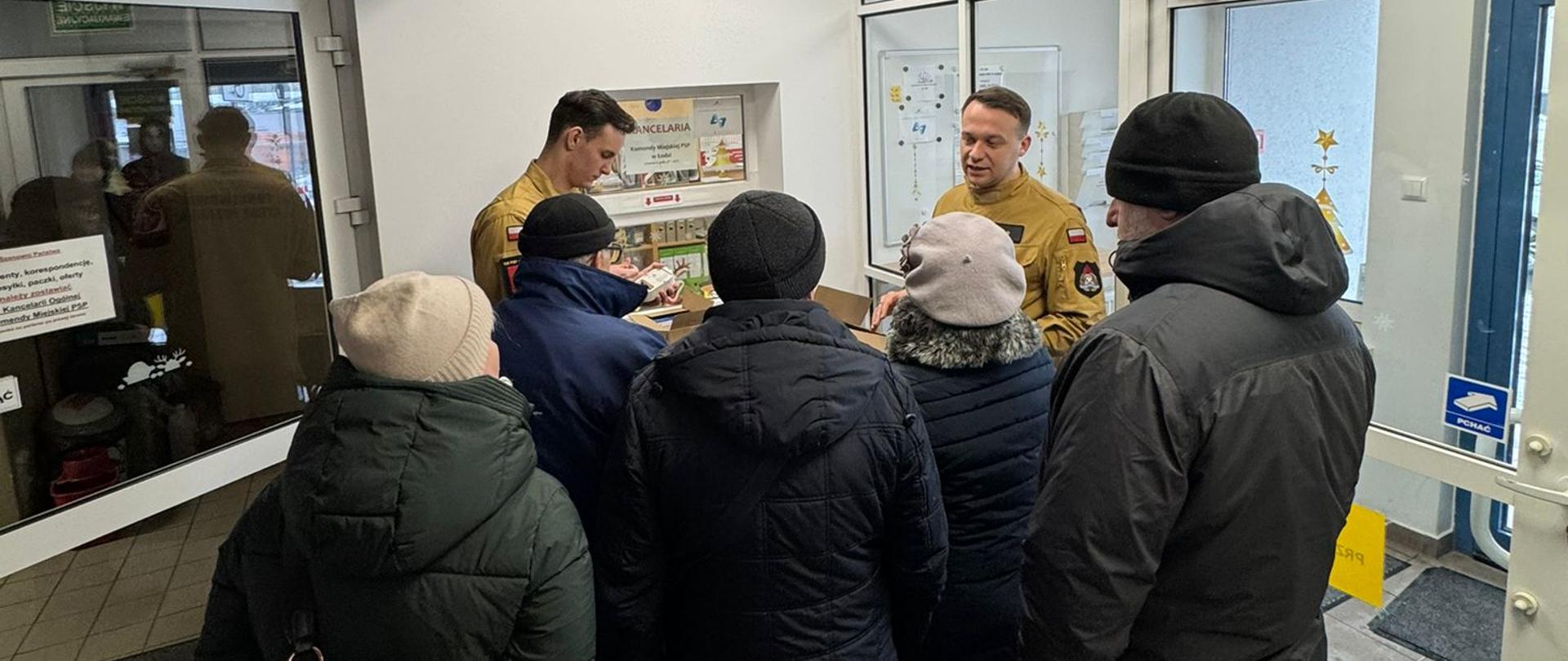 Dwóch strażaków w ubraniach służbowych przekazuje instrukcje montażu czujek osobą które zgłosiły się po ich odbiór