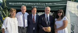 Na zdjęciu widać pięć osób stojących przed zielonym namiotem z napisem „Wojewódzki Inspektorat Ochrony Roślin i Nasiennictwa w Warszawie”. Obok namiotu znajduje się baner z logo instytucji. Jedna z osób trzyma bochenek chleba.