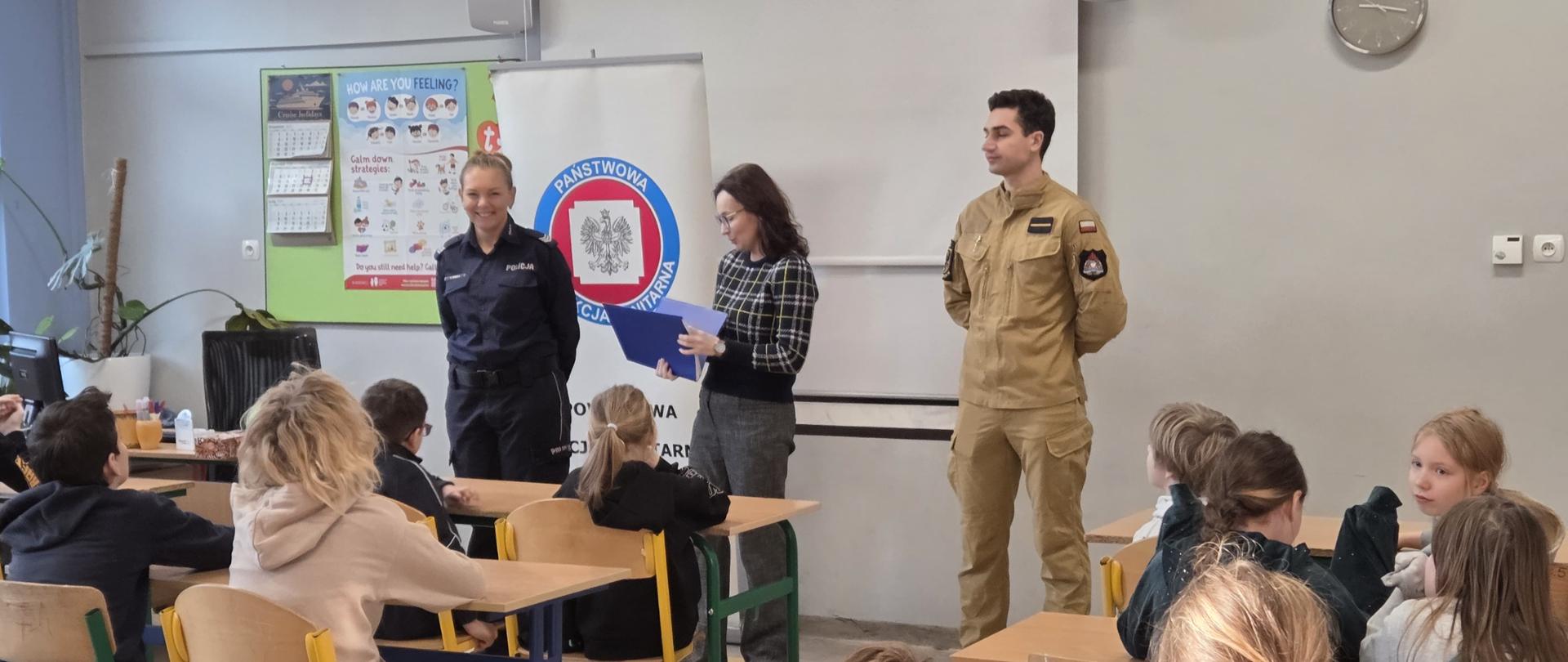 Zdjęcie przedstawia strażaków oraz policje przed dziećmi w szkole podstawowej