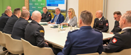 Zdjęcie przedstawia oficjalne spotkanie w sali konferencyjnej, w którym uczestniczą strażacy w mundurach oraz osoby cywilne siedzące przy wspólnym stole. W tle widoczne są banery informacyjne, a u dołu kadru umieszczono oficjalny pasek z logotypami Funduszy Europejskich, flagi Polski oraz Unii Europejskiej z napisem o dofinansowaniu.