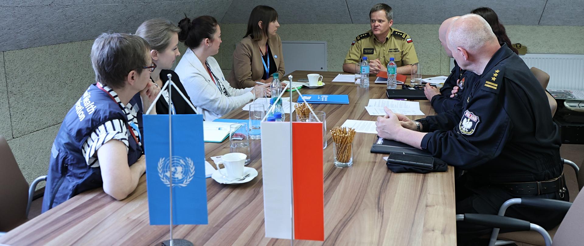 2 funkcjonariuszy KG PSP oraz 4 kobiety (po lewej stronie stołu) rozmawiają przy stole. Na stole stoją flagi WHO oraz Polski