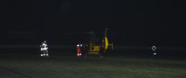 Śmigłowiec LPR na stadionie w Rawiczu. Maszyna w barwach żółtej i czerwonej stojąc na płycie boiska piłkarskiego przygotowuje się do odlotu z poszkodowaną. Przy śmigłowcu członek załogi, wokół - strażacy zabezpieczający start. Na dworze ciemno.