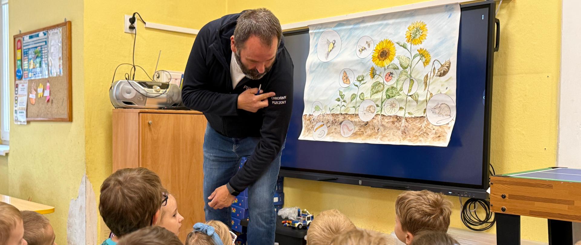 Inspektor prowadzi zajęcia edukacyjne dla grupy dzieci siedzących na podłodze w sali lekcyjnej. Obok niego znajduje się ekran z ilustracją przedstawiającą słoneczniki oraz owady i etapy ich rozwoju