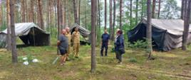 Na zdjęciu widzimy sześć osób stojących w lesie na terenie obozu harcerskiego. Widać też rozłożone namioty pomiędzy drzewami.