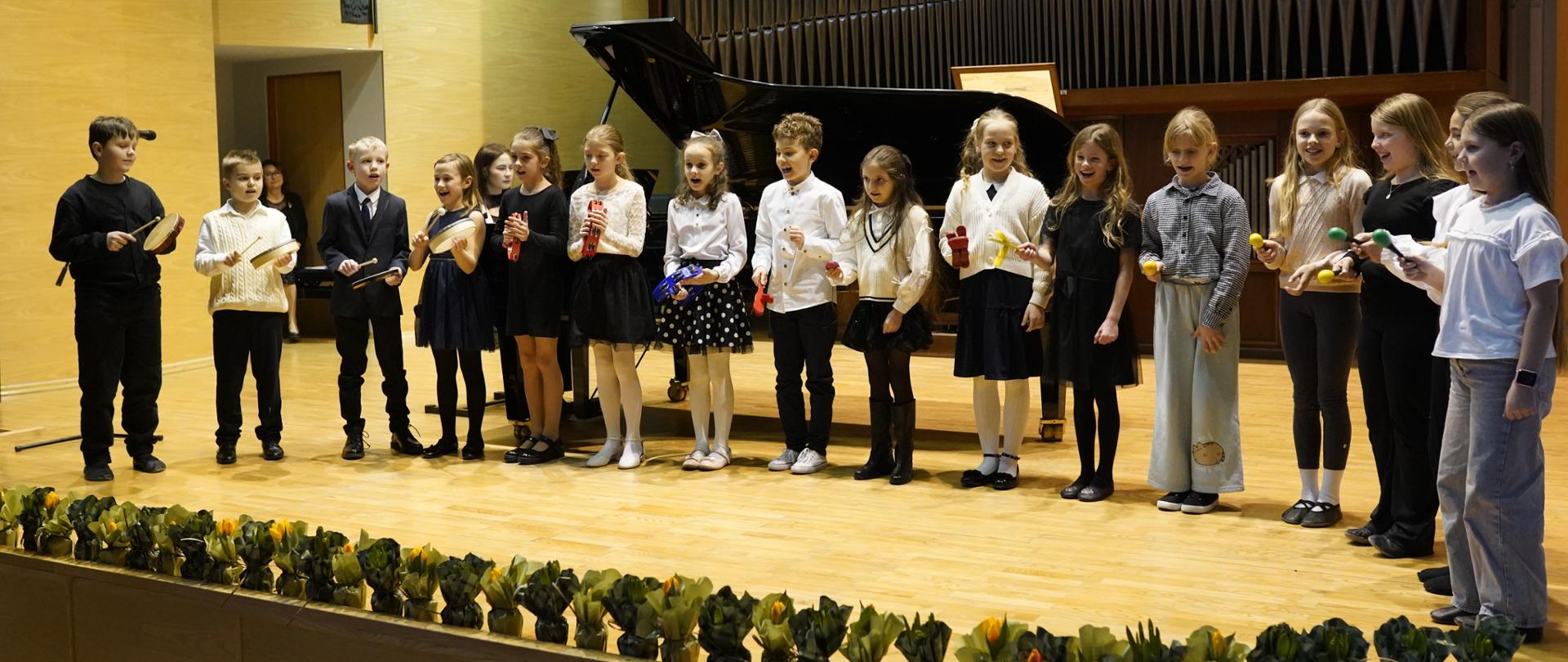 To długa scena w sali koncertowej, widziana z perspektywy widza na widowni.
Na środku sceny stoi rząd kilkunastu dzieci, ubrane w dość eleganckie stroje — przewaga jasnych koszul i ciemnych spódnic albo spodni; kilka dziewczynek ma sukienki z tiulem albo z wzorami. Wszyscy stoją bok w bok, zwróceni w stronę widowni. Dzieci różnią się wzrostem i wiekiem, ale wyglądają na wczesno i starsze klasy szkoły podstawowej. Trzymają w dłoniach kolorowe instrumenty perkusyjne, najprawdopodobniej marakasy lub inne małe grzechotki, w żywych barwach: żółte, czerwone, niebieskie, zielone. W tle, po lewej stronie sceny, stoi otwarty fortepian koncertowy ze schowanym wiekiem, czarny, o charakterystycznej dużej, poziomej obudowie — typowym dla fortepianu półkowego lub koncertowego; słowo grand piano oznacza fortepian o poziomej ramie i strunach. Za fortepianem, w głębi, widać dużą ścianę z pionowymi piszczałkami organów lub elementami ozdobnymi przypominającymi piszczałki; tworzą one wysoki, symetryczny wzór, sięgający niemal sufitu sali. Scena jest wykonana z jasnego drewna, dobrze oświetlona ciepłym, żółtawym światłem, które tworzy miękkie cienie. Podłoga sceny jest pusta poza dziećmi, fortepianem i dekoracją na przednim brzegu. Przed sceną, w dolnej części kadru, widać dekorację z regularnie rozmieszczonych małych zielonych roślin lub sztucznych dekoracji, które biegną wzdłuż całej szerokości frontu sceny. Po prawej stronie, nieco z tyłu w rzędzie, stoi kilka dzieci w bardziej casualowych ubraniach, ale nadal schludnych. Widać ich naturalne, skupione wyrazy twarzy, jakby właśnie występowali lub dopiero co skończyli grać. Scena sprawia wrażenie uroczystości szkolnej, występu muzycznego lub koncertu dziecięcego, w sali z profesjonalnym instrumentem i dużą publicznością.
