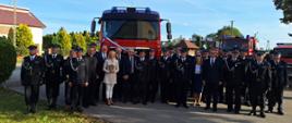 Zdjęcie grupowe przed przekazanym samochodem strażackim stoją wszyscy uczestnicy uroczystości, strażacy ubrani są w mundury galowe na drugim planie widać samochody strażackie stojące na parkingu