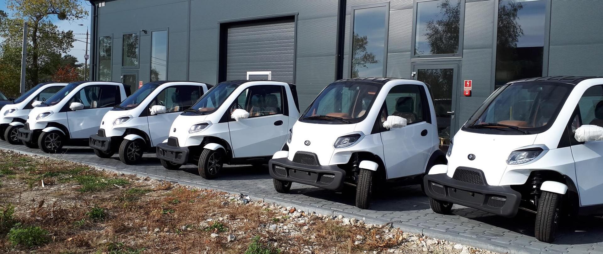 Rząd sześciu biało-czarnych aut przed halą przemysłową