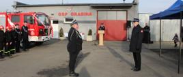 Zdjęcie przedstawia strażaków podczas składania meldunku o zakończeniu uroczystości. W tle budynek OSP Grabina, samochód pożarniczy oraz inni strażacy