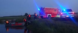 Pojazdy straży pożarnej stoją na brzegu rzeki. Strażacy wodują łódź