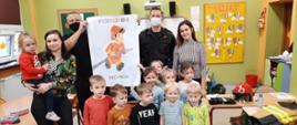 Obraz przedstawia zdjęcie zbiorowe, na którym znajdują się dzieci oraz dwóch strażaków. Zdjęcie ma miejsce w pomieszczeniu przedszkola, zielone ściany, piaskowo-zielone mebelki na których znajdują się zabawki oraz klocki. Strażacy ubrani w czarne ubrania sztabowe. Strażak stojący z lewej strony kadru trzyma w rękach kartkę z namalowanym strażakiem.