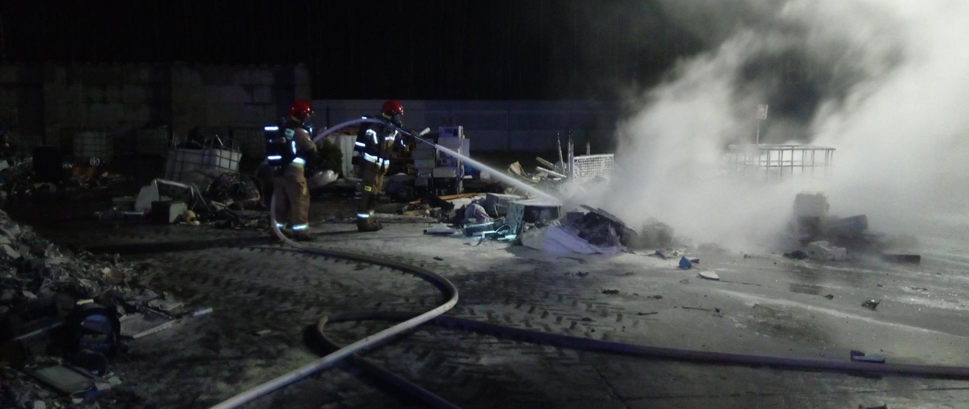 Zdjęcie przedstawia dwóch ratowników podczas gaszenia pryzmy urządzeń elektronicznych przeznaczonych do przetworzenia na placu składowym zakładu recyklingu.