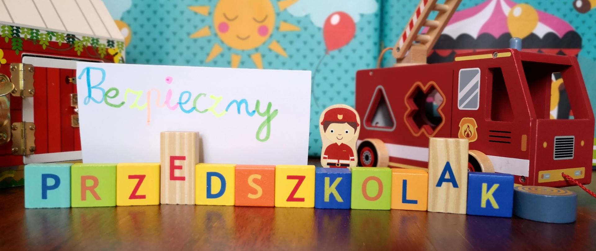 Na pierwszym planie umieszczono zapisany na kartce kolorowymi mazakami napis Bezpieczny, poniżej pod tym napisem z klocków z nadrukowanymi literami ułożono napis Przedszkolak. Za napisem stoi klocek w kształcie strażaka oraz drewniany wóz strażacki, za nimi w tle niebieska plansza ze słońcem i chmurami. Zabawki leżą na drewnianej podłodze.