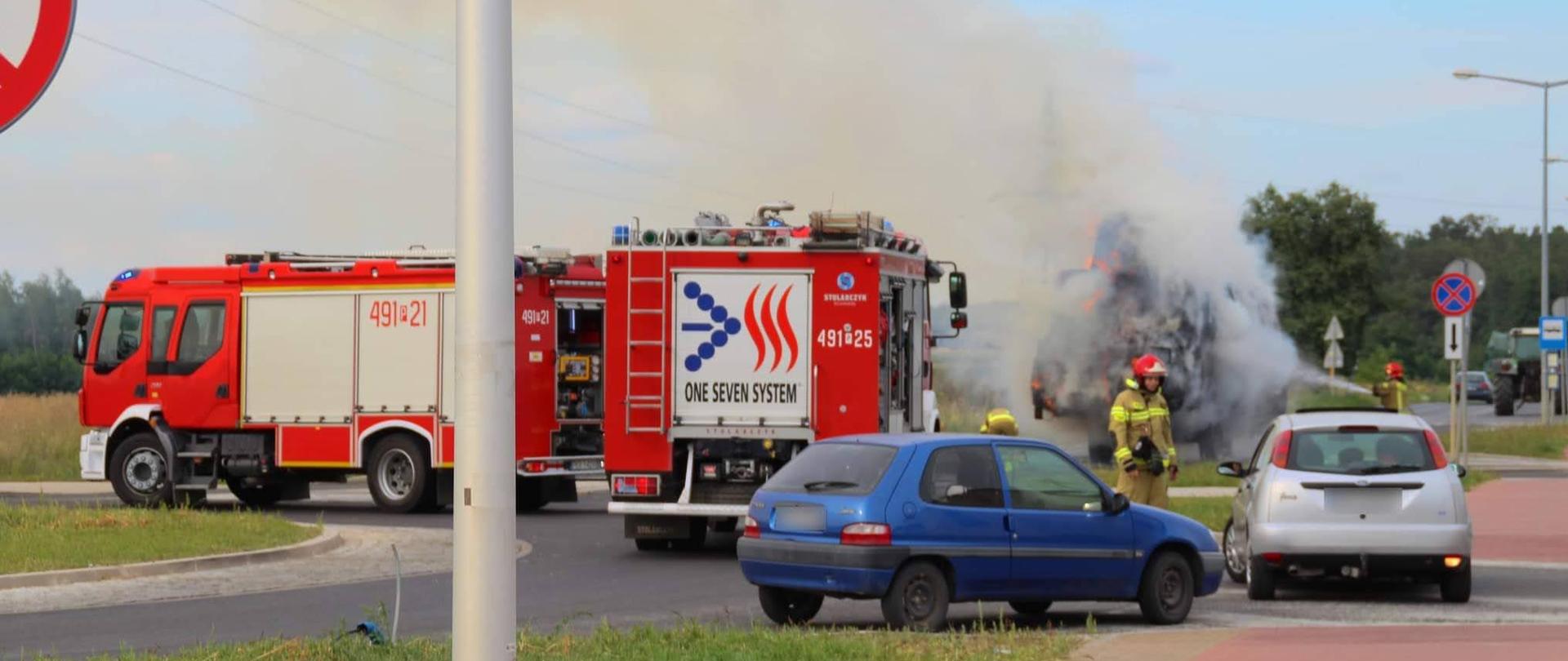 Pali się słoma w balotach na przyczepie rolniczej. Strażacy prowadzą działania gaśnicze. Na skrzyżowaniu o ruchu okrężnym, stoją dwa wozy strażackie blokując drogę. W oddali widać strażaka który podaje prąd wody celem ugaszenia pożaru. Inny strażak informuje ludzi o braku możliwości przejazdu drogą.