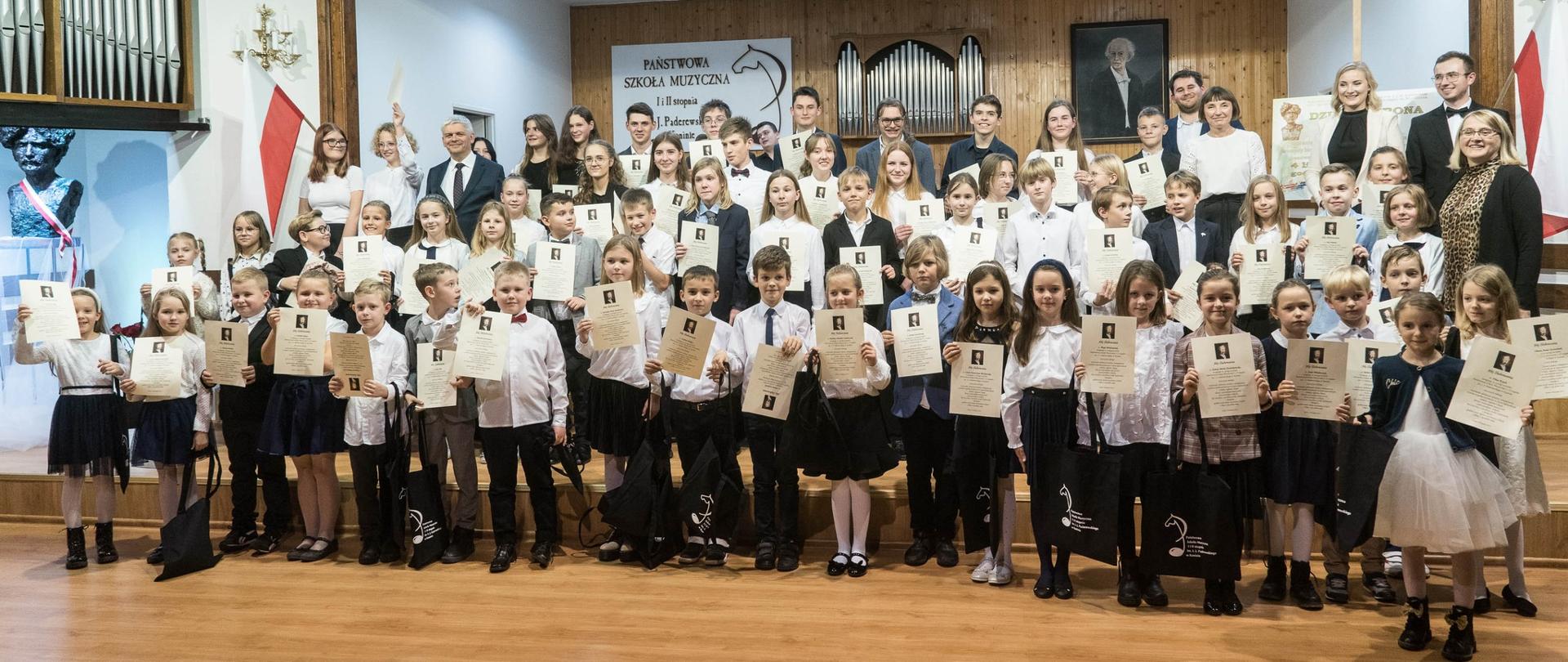 Na scenie PSM stoją wszyscy uczniowie klas pierwszych w towarzystwie dyrekcji i nauczycieli ubrani w stroje galowe. W ręku trzymają akty ślubowania i pamiątkowe torby.