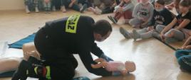 Strażak klęczy i trzyma na ręku manekina niemowlęcia. Wkoło na podłodze siedzą dzieci.