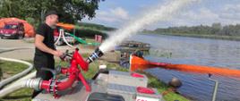 Na zdjęciu widoczne stojące tuż przy nabrzeżu, na trawniku działko gaśnicze w kolorze stalowym na kołach, z którego wyrzucany jest strumień rozproszonej wody, sterowane przez strażaka w czarnych długich spodniach mundurowych, krótkiej czarnej koszulce i czapce na głowie. Za nim przyczepka z wieloma podłączonymi wężami białymi, zielonymi. Obok przód stojącego czerwonego samochodu osobowego, za którym widać pomarańczowy namiot pneumatyczny. W tle drzewa, zbiornik wodny z widocznym fragmentem pomarańczowego odcinka zapory i błękitne niebo z niewielkimi obłokami.