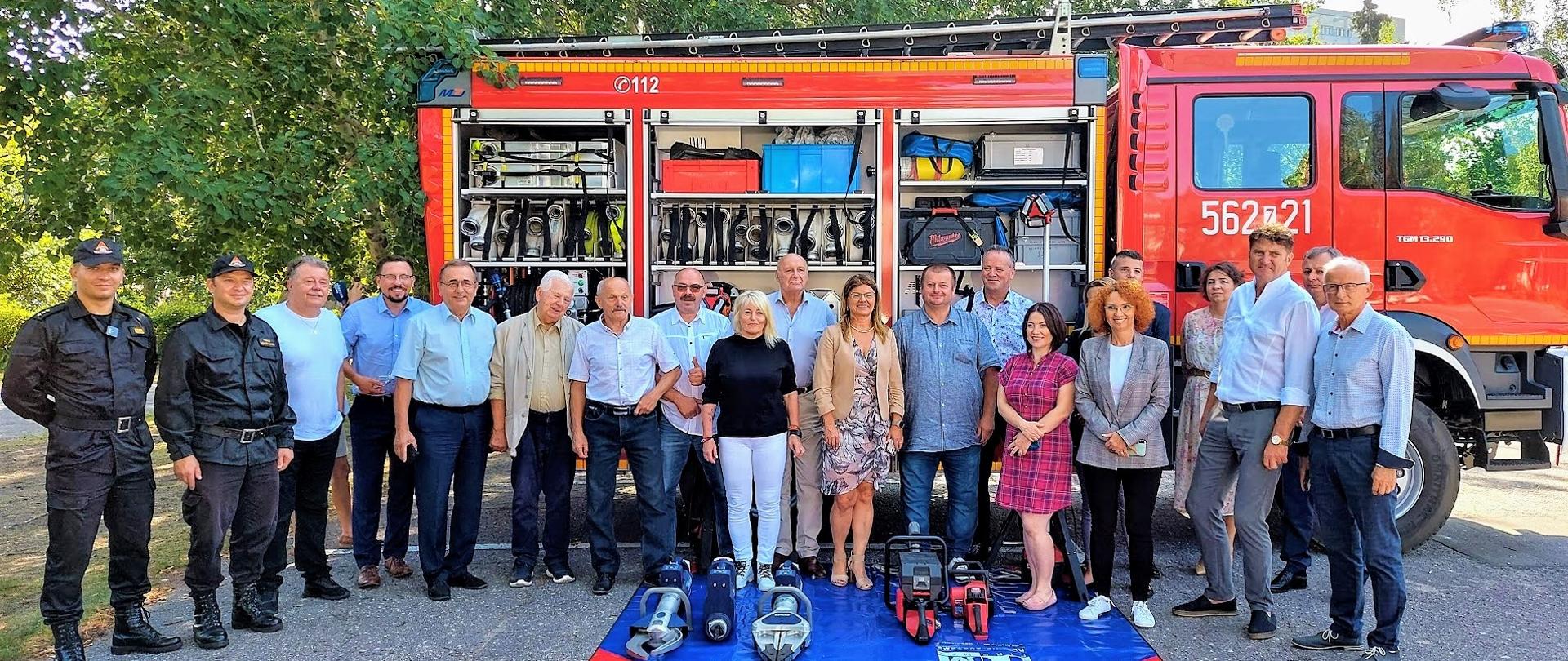 Uczestnicy sesji Rady Miasta wraz z funkcjonariuszami Komendy Miejskiej Państwowej Straży Pożarnej w Świnoujściu. 21 osób na tle pojazdu pożarniczego z wyeksponowanym sprzętem ratowniczym. Przed stojącymi mata sprzętowa z narzędziami ratownictwa technicznego.