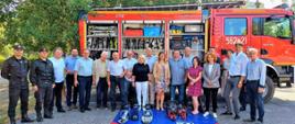 Uczestnicy sesji Rady Miasta wraz z funkcjonariuszami Komendy Miejskiej Państwowej Straży Pożarnej w Świnoujściu. 21 osób na tle pojazdu pożarniczego z wyeksponowanym sprzętem ratowniczym. Przed stojącymi mata sprzętowa z narzędziami ratownictwa technicznego.