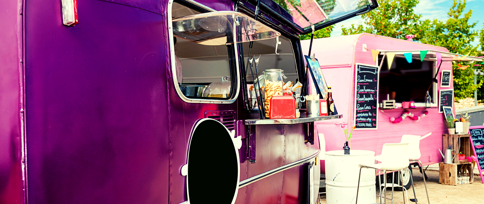 Dwa food trucki - przyczepy, z których serwowane jest jedzenie. Jedna przyczepa (po lewej) - fioletowa, druga (po prawej) - różowa. Widoczne stojące na blatach przystawki i napoje. Przed przyczepami krzesła.