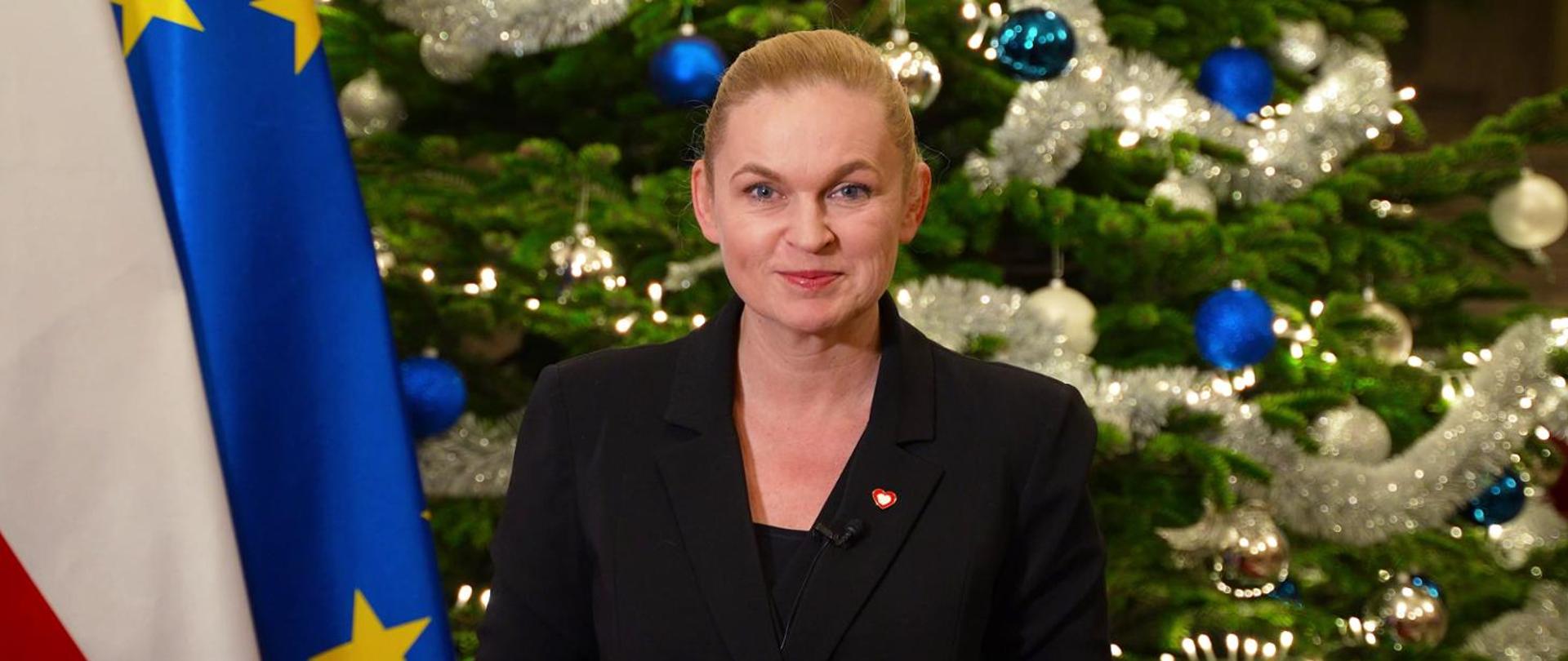 Ministra Barbara Nowacka stoi na tle udekorowanej bombkami choinki.