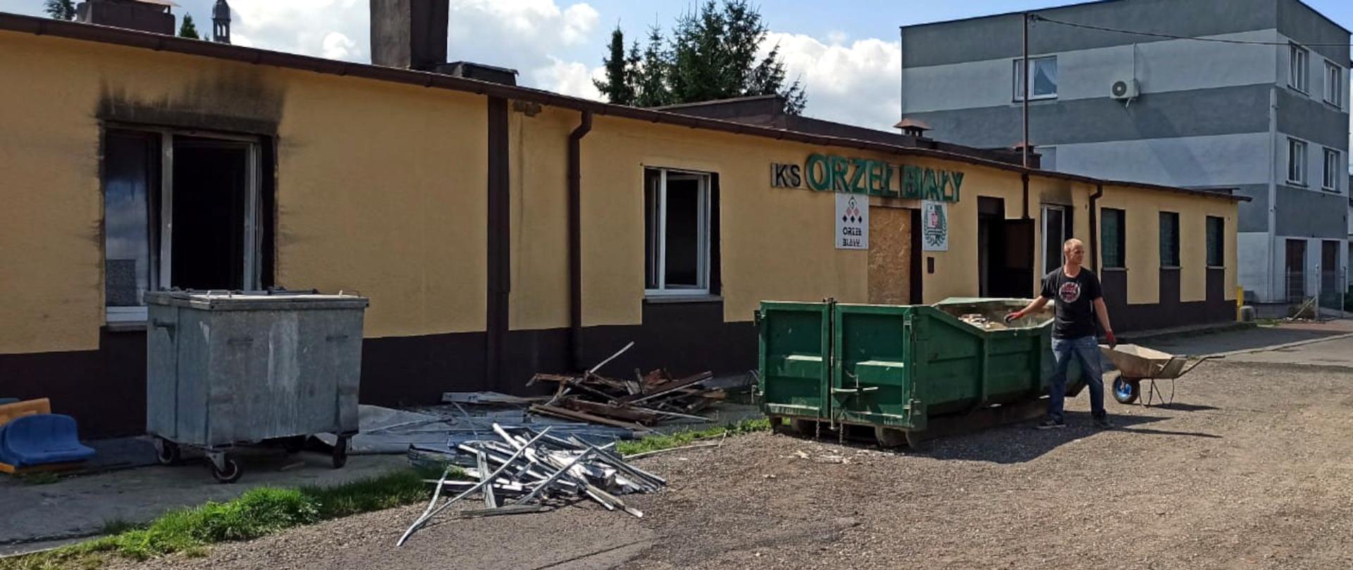 Zdjęcie przedstawia częściowo zniszczoną elewację budynku administracyjnego klubu sportowego Orzeł Biały. Przed budynkiem widać kontenery na gruz.