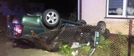 Zdjęcie przedstawia samochód osobowy leżący na dachu przy budynku mieszkalnym jednorodzinnym. Samochód uszkodził ogrodzenie posesji. Pora nocna. 