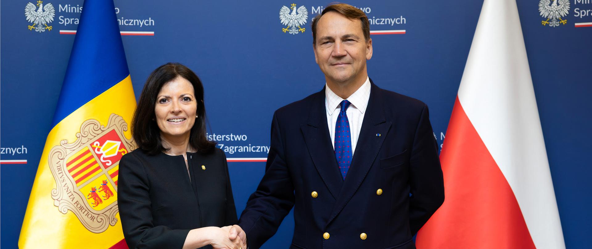 Foreign ministers of Poland and Andorra A woman and a man in formal attire shake hands. The woman has shoulder-length dark hair and is wearing a black blazer with a black-and-white patterned skirt. The man is dressed in a navy double-breasted blazer with gold buttons, a white shirt, and a blue tie. In the background are the flags of Andorra and Poland, along with a blue backdrop displaying the emblem and name of the Ministry of Foreign Affairs.