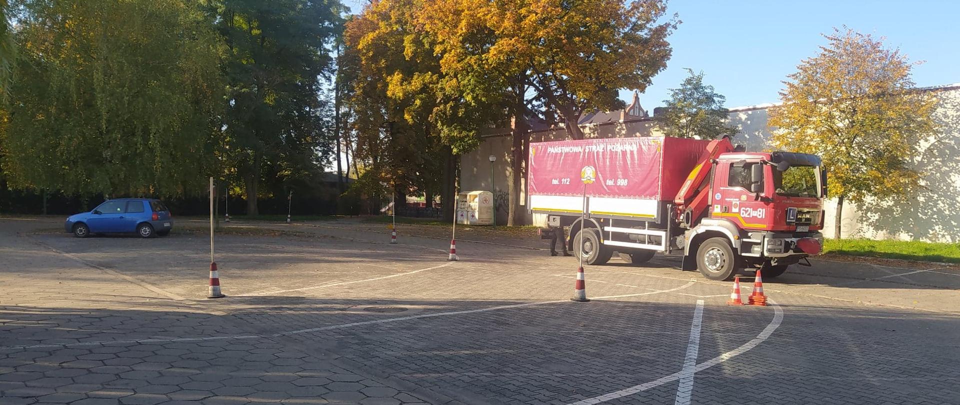 Szkolenie kierowców. Plac manewrowy na terenie targowiska miejskiego w Rawiczu. Na placu stoi samochód strażacki - ciężarowy, na którym odbywa się praktyczna nauka jazdy. Przed samochodem rozstawione są pachołki i tyczki oraz wymalowane białe poziome linie - wyznaczające tory jazdy. W tle budynek, drzewa oraz samochód osobowy.