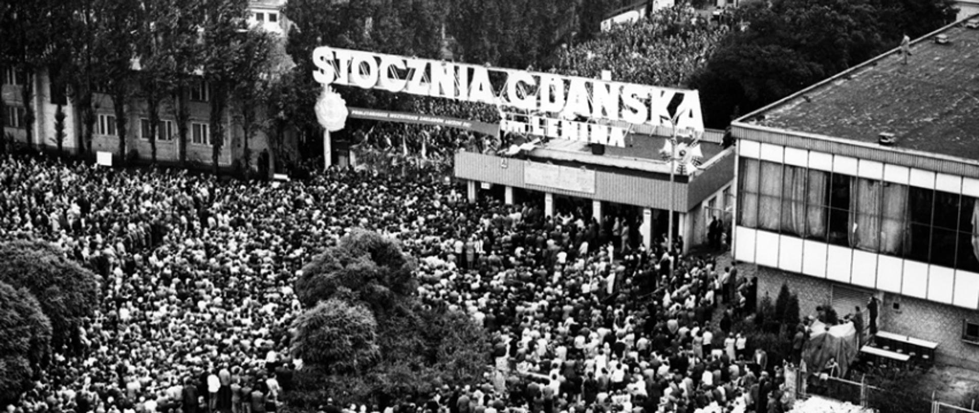August strike at Lenin's Shipyard in Gdansk. Top view of Gate No. 2 and the citizens and shipyard workers gathered below it. Above the gate, there is a banner with the slogan "PROLETARIANS OF ALL FACTORIES UNITE!", and on the roof of the guardhouse there are boards with 21 demands of MKS.