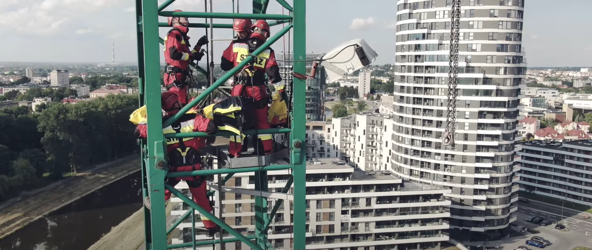 Na zdjęciu ratownicy grupy wysokościowej w umundurowaniu podczas ćwiczeń przy użyciu technik alpinistycznych ewakuują poszkowanego z żurawia budowlanego. W tle zabudowa miejska miasta Rzeszowa oraz rzeka Wisłok.