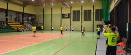 Zawodnicy rywalizujący podczas meczu futsalu w hali sportowej – drużyny w żółtych i białych strojach są w trakcie dynamicznej akcji.