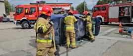 Na placu leży przewrócony na bok granatowy samochód osobowy, obok strażacy ubrani w ubrania specjalne prowadza pokazowa akcje ratownicza. Z tyłu widać czerwone wozy bojowe straży pożarnej.