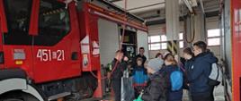 Grupa dzieci ze Szkoły Podstawowej nr 3 w Łowiczu oglądają ubrania żaroodporne prezentowane przez strażaków w tle samochód ratowniczo gaśniczy barwy czerwonej z numerem bocznym na drzwiach 451 E 21 