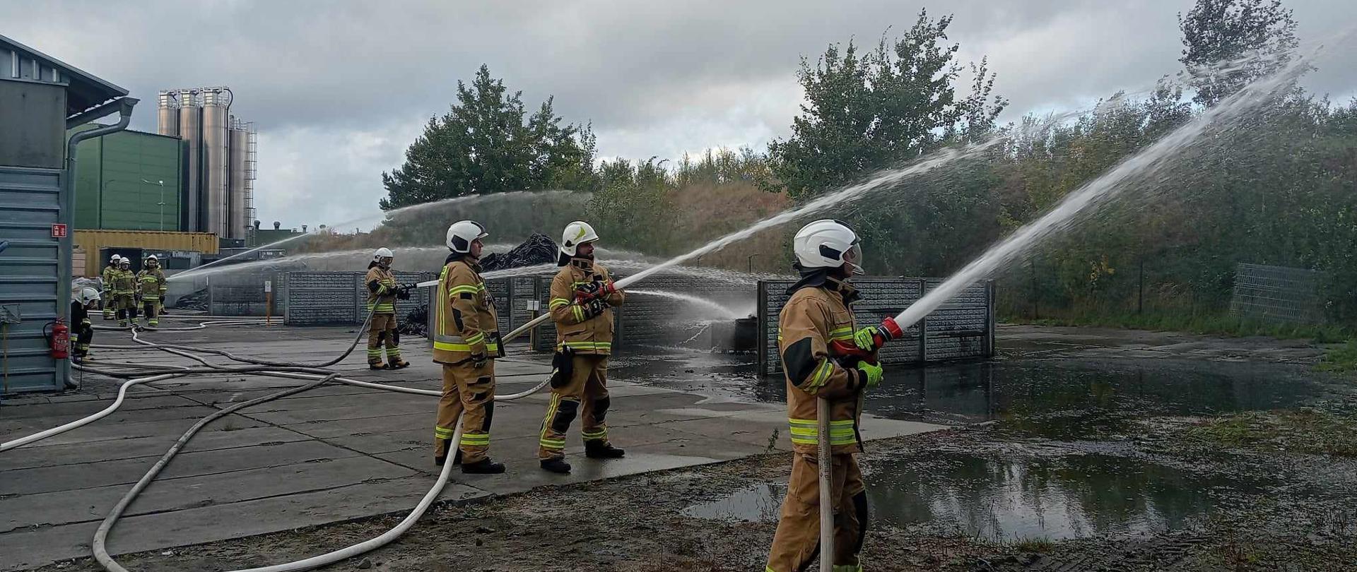 Zdjęcie przedstawia strażaków podczas ćwiczeń taktyczno – bojowych na terenie zakładu produkującego opony i dętki.
W tle budynki i teren zakładu.
