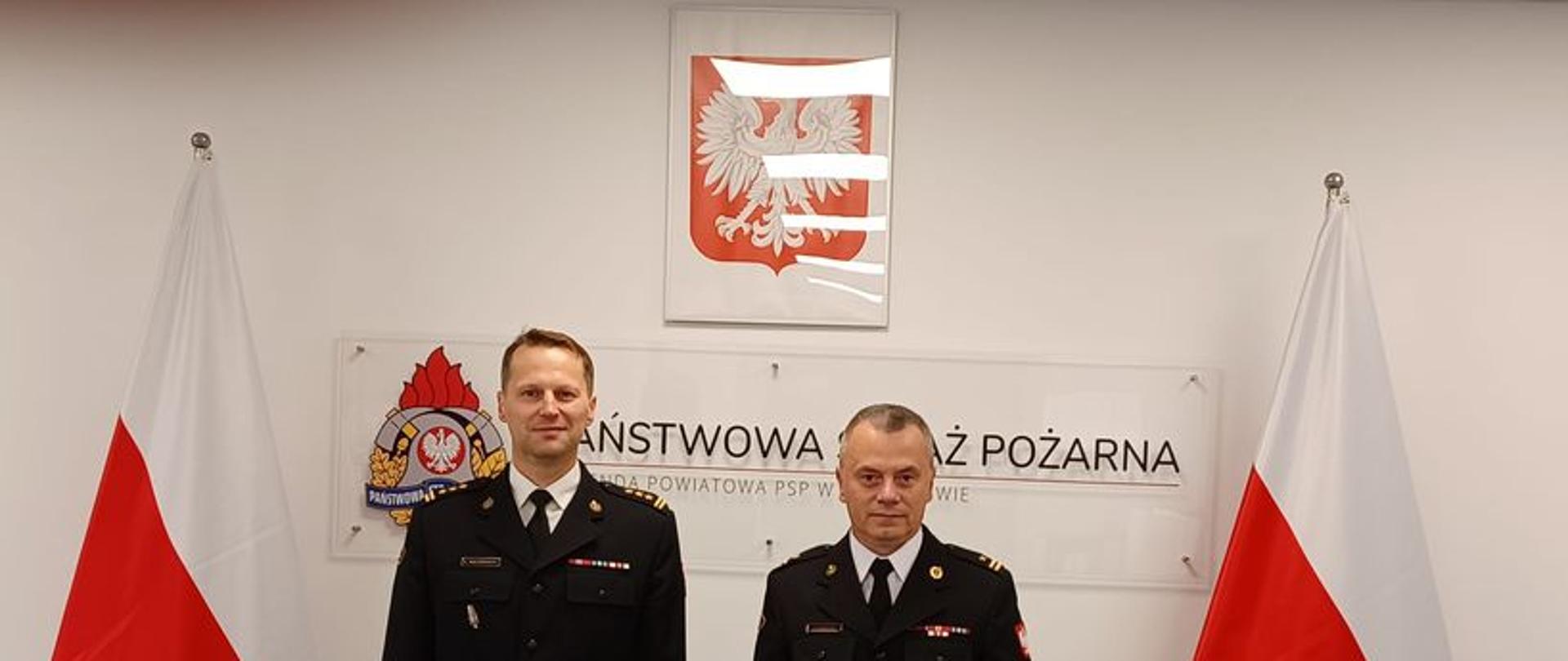 Komendant Powiatowy PSP w Człuchowie wraz z mianowanym Dowódcą Jednostki Ratowniczo-Gaśniczej stojący pomiędzy flagami Polski, w tle logo z nazwą Komendy Powiatowej PSP.