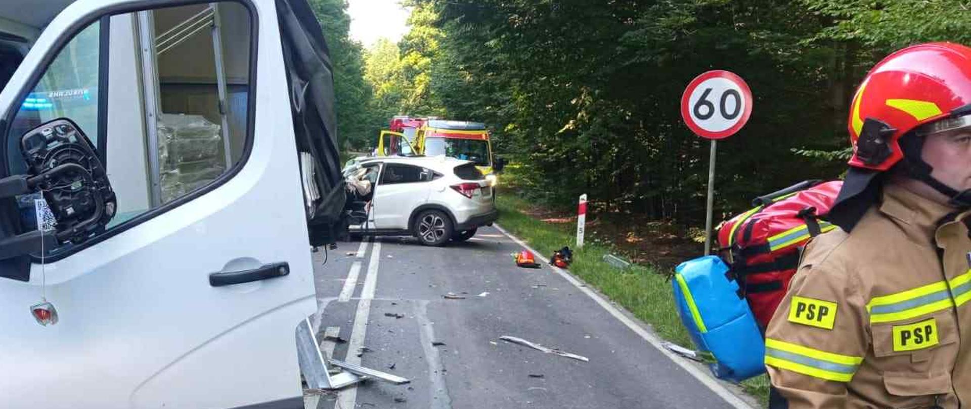 Na zdjęciu widać uszkodzone samochody: osobowy oraz dostawczy. Na jezdni są rozrzucone części pojazdów. Strażak PSP idzie z torbą medyczną.