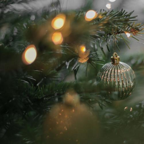 Niewyraźne zdjęcie gałązki choinkowej. Na niej wyostrzony obraz zawieszonej okrągłej bombki koloru złotosrebrnego. Widoczne są również rozmyte światełka.
