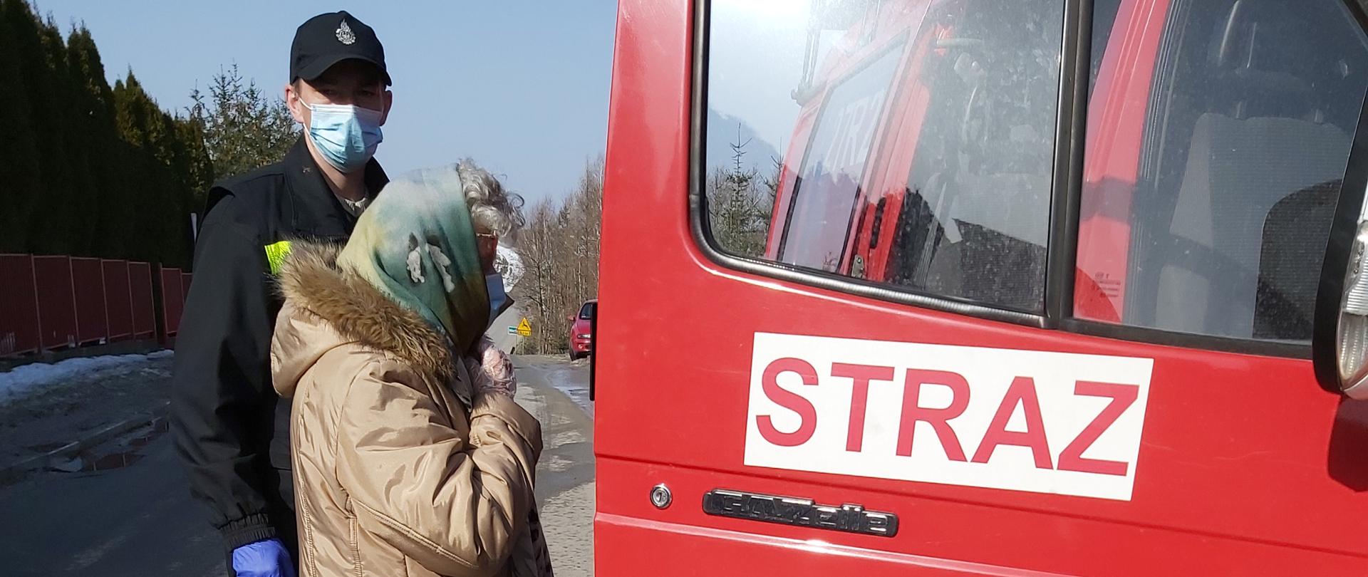 Na zdjęciu widnieje strażak w umundurowaniu koszarowym, rękawiczkach i masce oraz kobieta, która udaje się na szczepienie. Znajdują się przed otwartymi drzwiami samochodu pożarniczego typu bus z zamiarem wejścia do środka pojazdu.