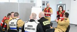 Strażacy siedzą przy stole podczas ćwiczenia dwóch funkcjonariuszy ubranych w czerwone kamizelki stoi przy tablicy i obserwuje.
