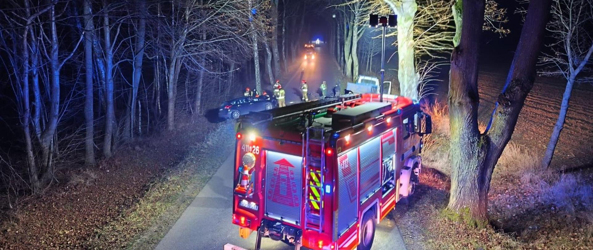 Zdjęcie zrobione nocą. Samochody straży pożarnej oświetlają drogę, wzdłuż drogi drzewa. Na drodze stoi duży czerwony samochód straży pożarnej dalej w tle dwa samochody które uczestniczyły w zdarzeniu oraz strażacy na drodze.
