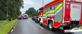 Na zdjęciu znajduje się samochód osobowy stojący na poboczu, samochód osobowy znajdujący się w rowie, w tle widać strażaków i karetkę pogotowia oraz wóz strażacki