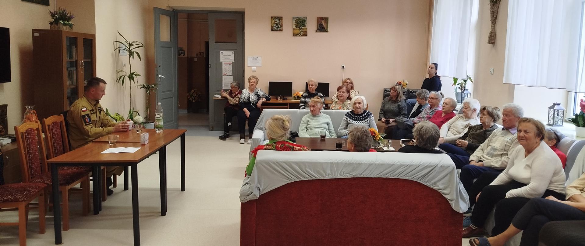 Na zdjęciu widać strażaka, który prowdzi rozmowy z osobami starszymi w pomieszczeniu budynku Domu Seniora. Na zdjęciu widać również seniorów siedziących przodem do strażaka na kanapach.