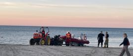 Pojazd mechaniczny wyciąga na ląd łódź ratowników, umocowaną na przyczepce transportującej. Nadzorują to funkcjonariusze. Całość odbywa się na brzegu plaży. W tle widać horyzont przed zmierzchem.