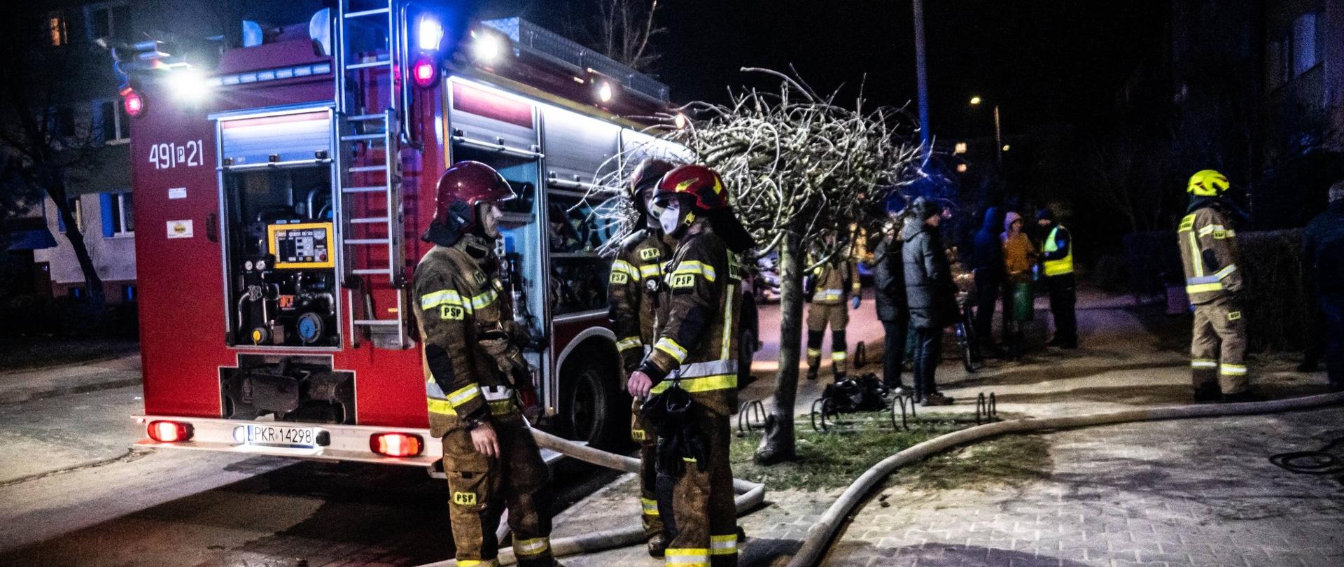 Zdjęcie przedstawia strażaków stojących przy pojeździe pożarniczym w trakcie realizacji zadań służbowych.