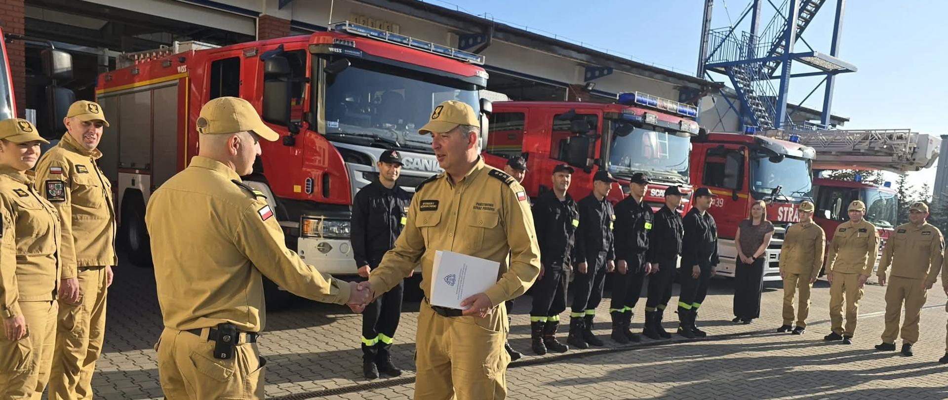 Na zdjęciu widać dwóch strażaków w beżowych mundurach, którzy podają sobie ręce. Jeden z nich trzyma w dłoni dokumenty. Obok nich stoi grupa innych strażaków, ubranych zarówno w beżowe, jak i czarne mundury. W tle widać trzy czerwone wozy strażackie z napisem "SCANIA" oraz "STRAŻ". Tło stanowią budynki remizy, a na horyzoncie widać również fragment niebieskiej, metalowej konstrukcji. Wszystkie osoby stoją na brukowanym placu w słoneczny dzień.